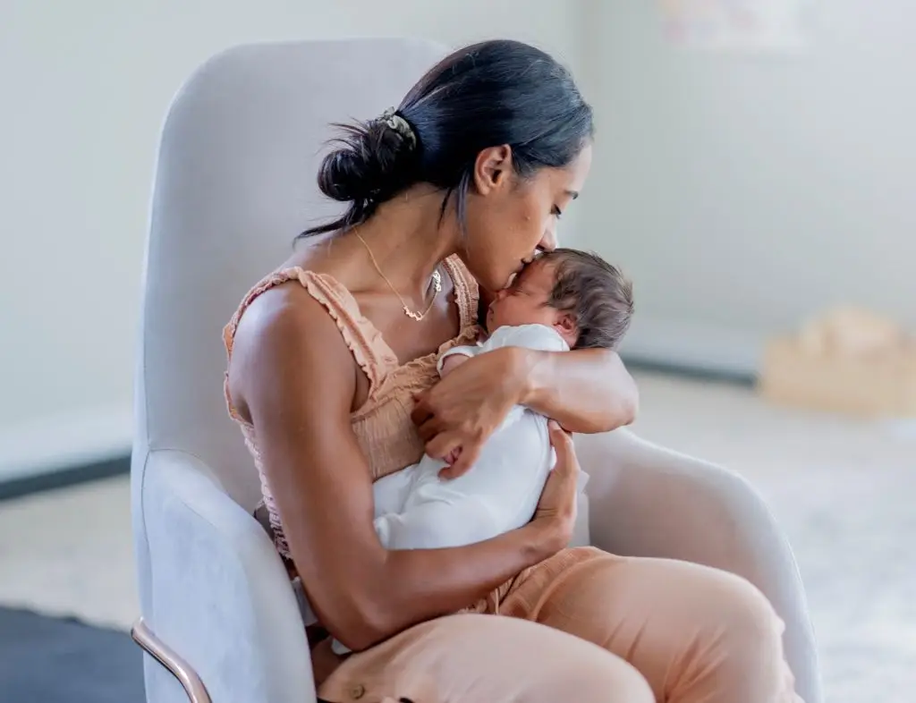 A new Mother sits in a rocking chair with her newborn cradled tightly in her arms as she kisses his forehead and bonds with her baby.